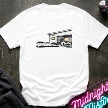 A white short-sleeve t-shirt laid flat on a dark textured surface, featuring a black and white graphic print of a garage with several cars in front. 