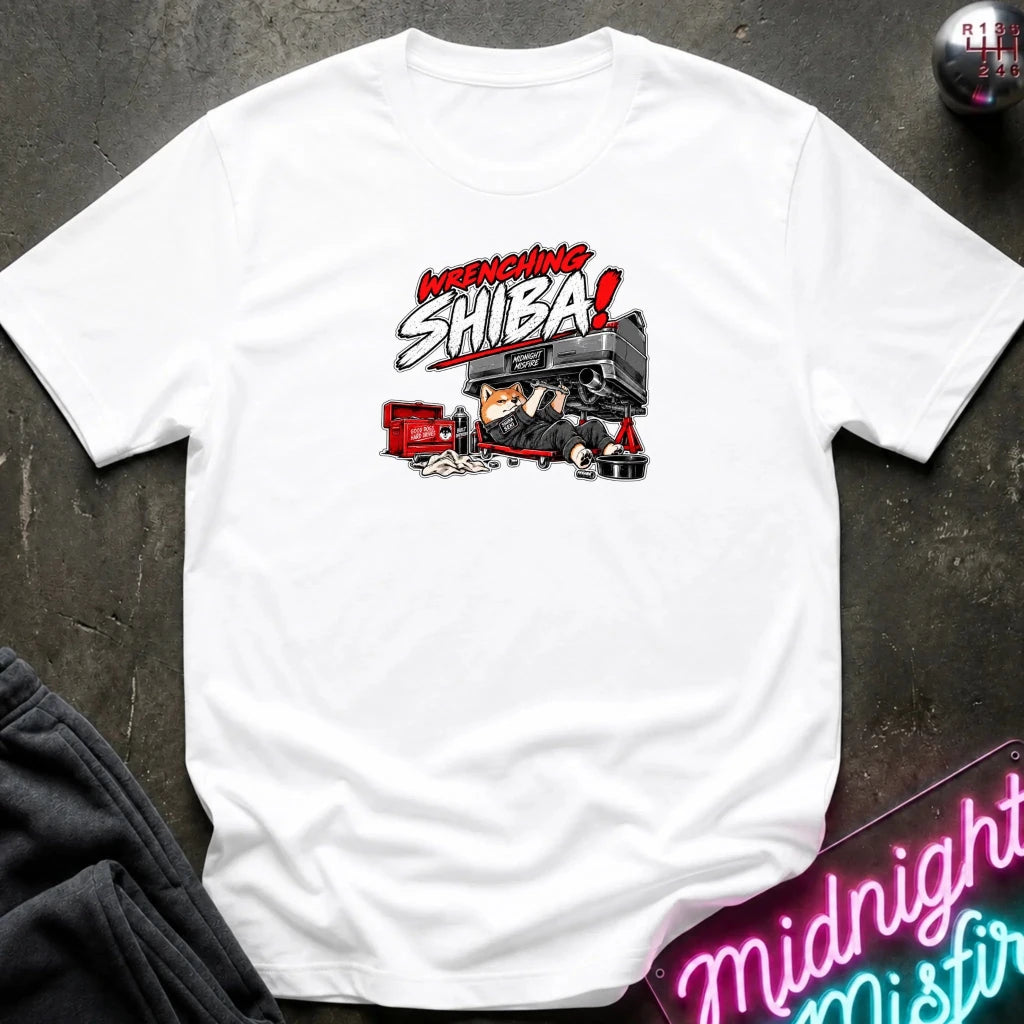 A white short-sleeve graphic t-shirt laid flat on a dark textured surface. The graphic features the text 'WRENCHING SHIBA!' in red and white comic-style lettering above an illustration of a character in a black and red vehicle.