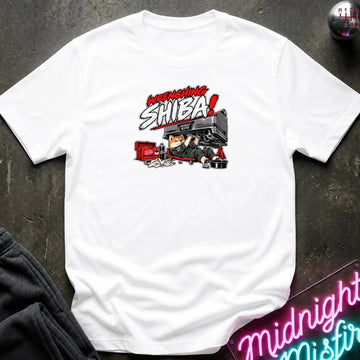A white short-sleeve graphic t-shirt laid flat on a dark textured surface. The graphic features the text 'WRENCHING SHIBA!' in red and white comic-style lettering above an illustration of a character in a black and red vehicle.