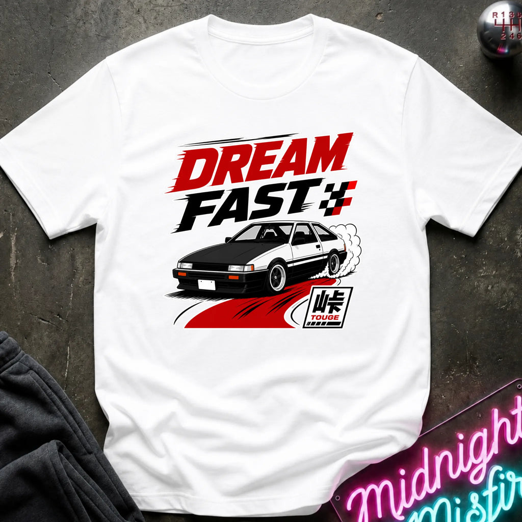A white short-sleeve graphic T-shirt laid flat on a dark textured surface. The shirt features a large front print with the words “DREAM FAST” in red and black, a black-and-white sports car, red motion lines, and a small Japanese-style logo.