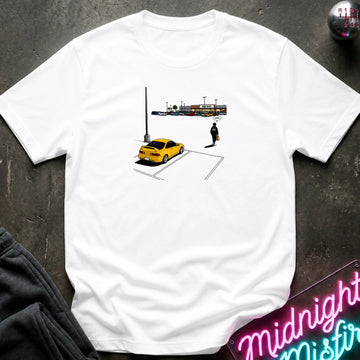 A white short-sleeve t-shirt laid flat on a dark textured surface, featuring a small graphic print of a yellow car, a parking lot, and a lone figure. 