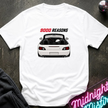 A white short-sleeve t-shirt laid flat on a dark textured surface, featuring a graphic of a white convertible sports car with the text '9000 REASONS' above it in red and black. A neon sign and dark pants are partially visible as props.