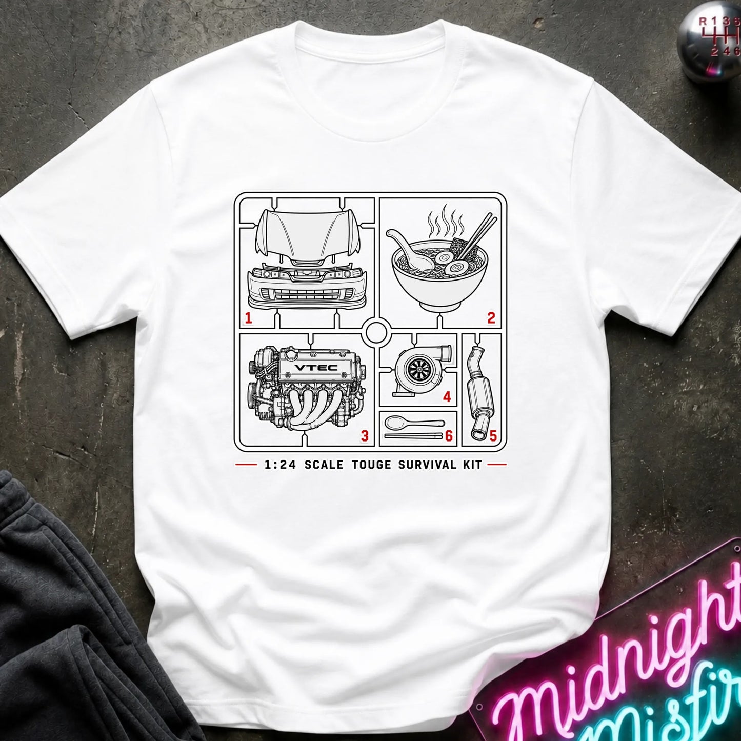 A white short-sleeve graphic T-shirt laid flat on a dark textured surface. The shirt features a black line-art schematic of a survival kit with numbered illustrations and the text '1:24 SCALE TOUGHE SURVIVAL KIT' printed on the chest.