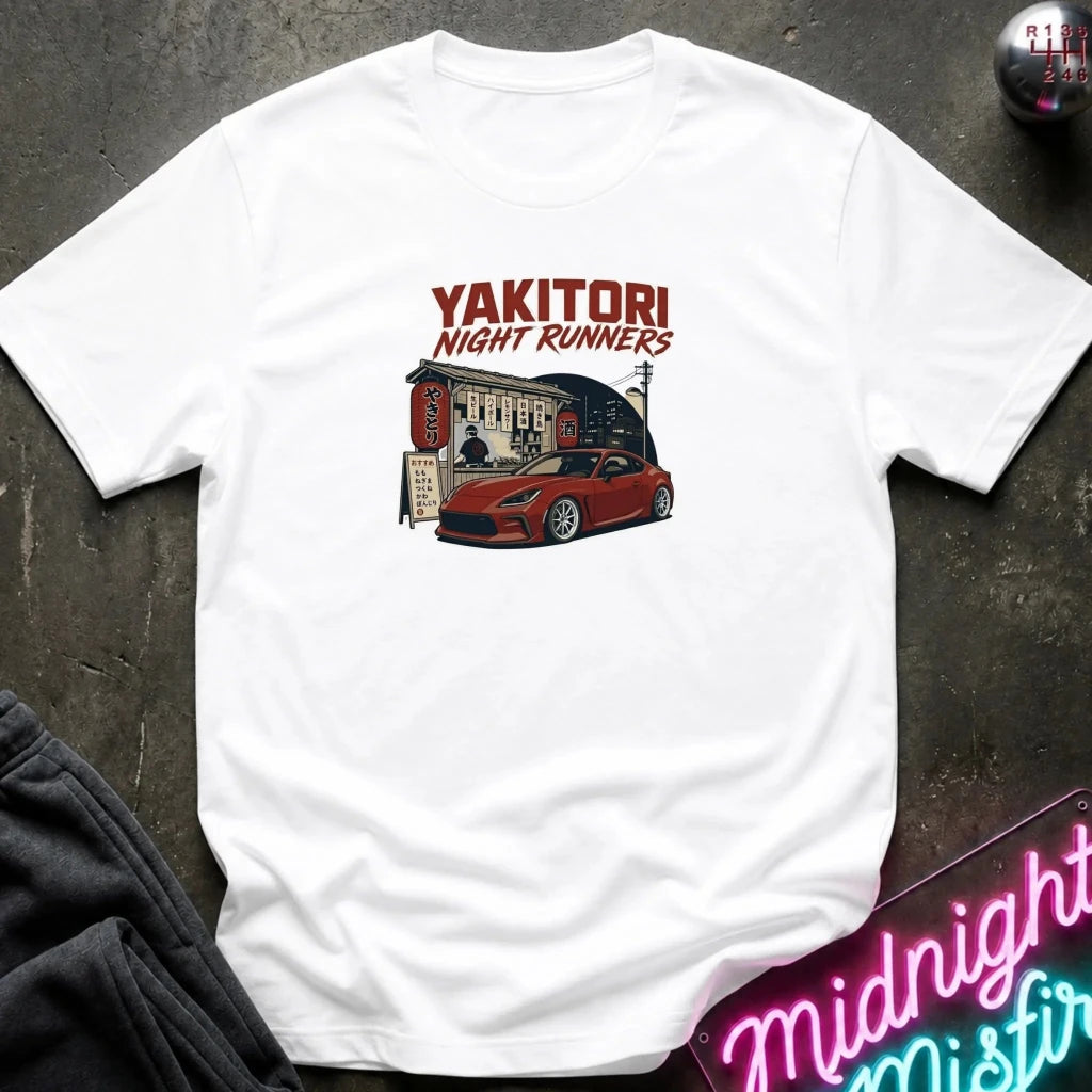 A white short-sleeve graphic T-shirt laid flat on a dark textured surface. The shirt features a red and black graphic print with the text 'YAKITORI NIGHT RUNNERS' above an illustration of a red sports car outside a restaurant.