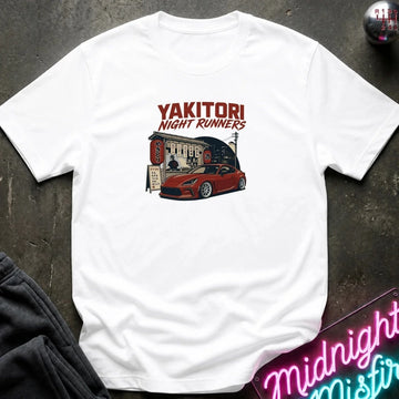 A white short-sleeve graphic T-shirt laid flat on a dark textured surface. The shirt features a red and black graphic print with the text 'YAKITORI NIGHT RUNNERS' above an illustration of a red sports car outside a restaurant.