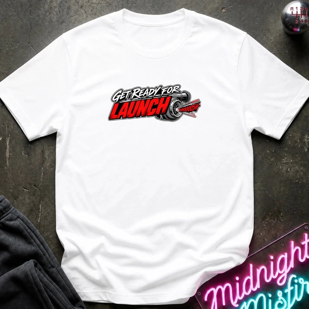 A white short-sleeve crewneck t-shirt laid flat on a dark textured surface. The shirt features a graphic on the chest that reads 'GET READY FOR LAUNCH' in stylized red, black, and white lettering, accompanied by a tire illustration.
