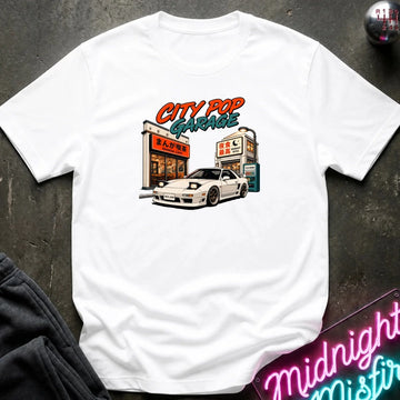 A white short-sleeve t-shirt laid flat on a dark textured surface, featuring a colorful retro-style graphic of a white sports car parked outside a garage with the text 'CITY POP GARAGE'.