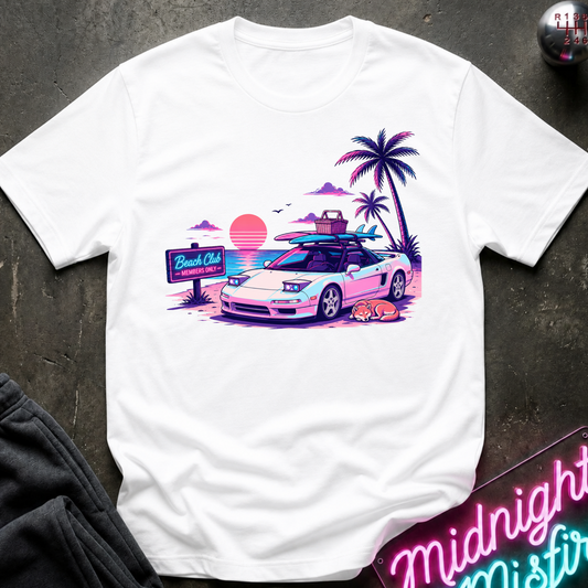 A white short-sleeve graphic t-shirt laid flat on a dark textured surface. The graphic features a retro-style illustration of a white sports car with surfboards on its roof, next to palm trees, a sunset, sleeping dog and a 'Beach Club' sign.