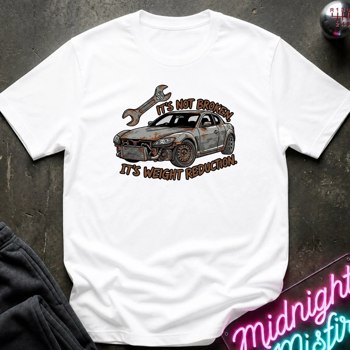 A white short-sleeve graphic T-shirt laid flat on a dark textured surface. The shirt features a distressed graphic of a damaged car with a wrench above it and the text 'IT'S NOT BROKEN IT'S WEIGHT REDUCTIVE' in brown.