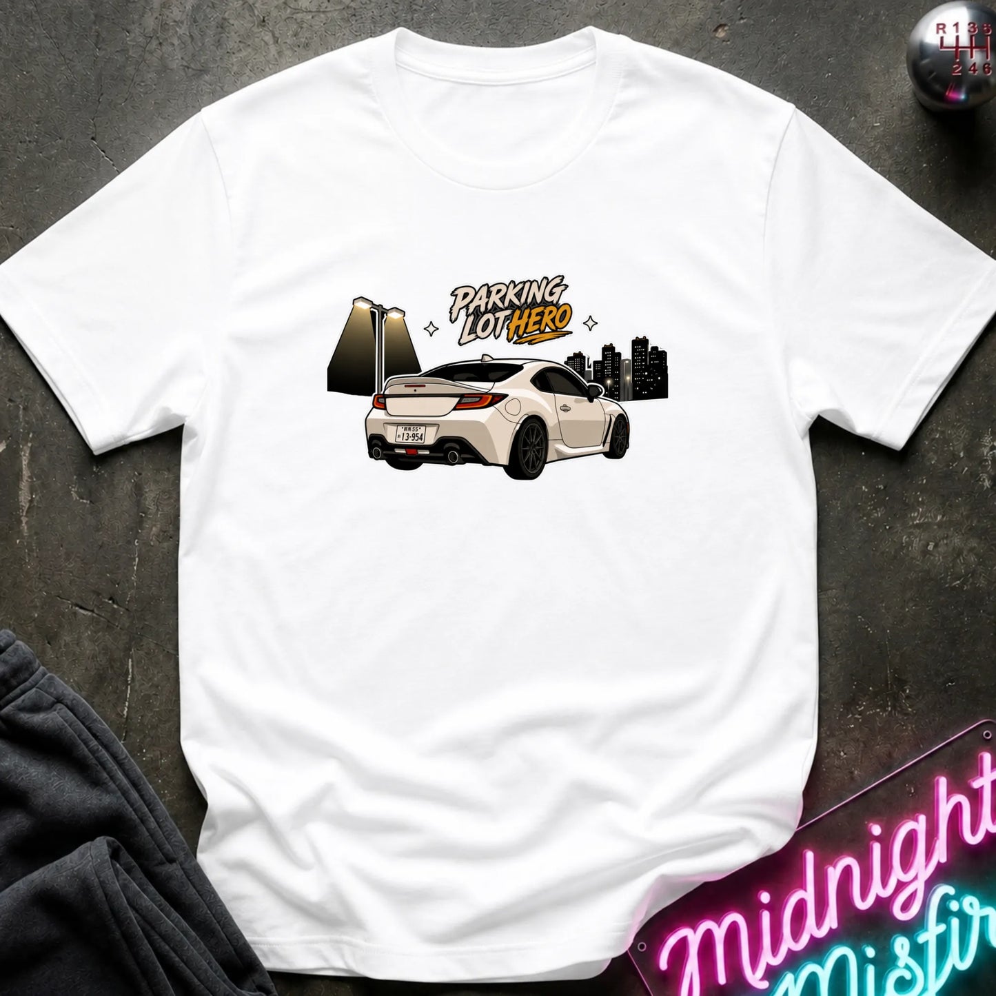 A white short-sleeve t-shirt laid flat on a dark textured surface, featuring a graphic print of a white sports car with the text 'PARKING LOT HERE' and a city skyline. A neon sign reading 'Midnight Misfire' is partially visible in the bottom right corner.
