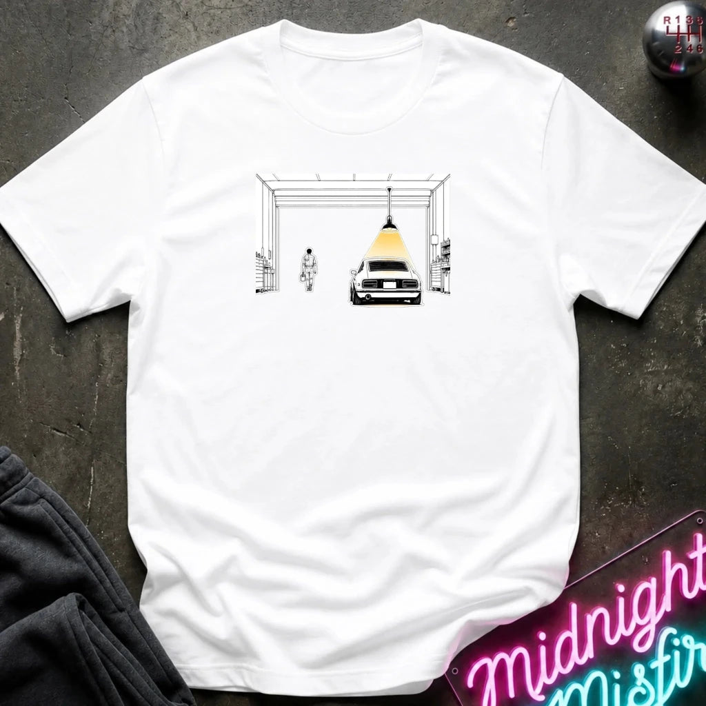 A white short-sleeve crewneck t-shirt laid flat on a dark textured surface. The center of the shirt features a graphic print of a vintage black car with a yellow dome light hanging beneath a white frame structure.