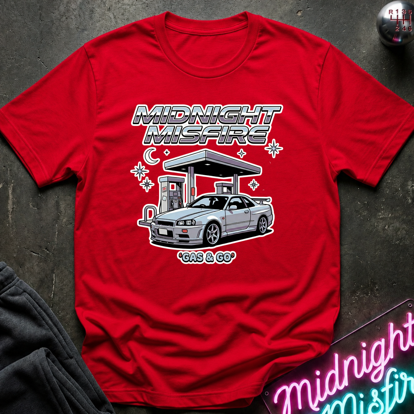 A red short-sleeve graphic T-shirt laid flat on a dark textured surface. The shirt features a large front print with the words “MIDNIGHT MISFIRE” above an illustration of a silver sports car at a gas station, with “GAS & GO” below.