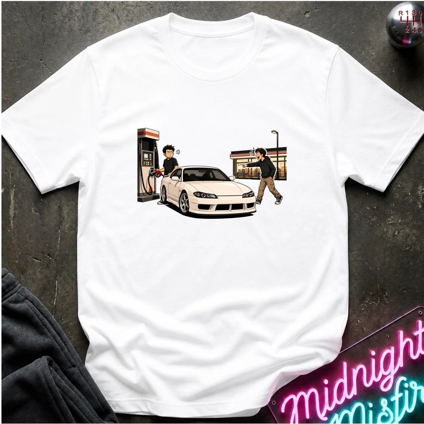 A white short-sleeve crewneck t-shirt laid flat on a dark textured surface. The shirt features a graphic print of a white sports car at a gas station with two characters, one pumping gas and the other walking away.
