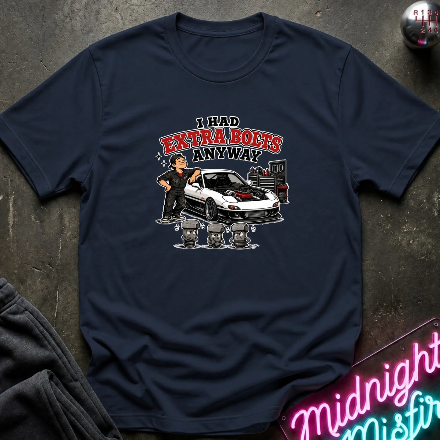 A navy blue short-sleeve graphic t-shirt laid flat on a dark textured surface. The graphic features a cartoon of a man standing next to a white sports car with red detailing, with the text 'I HAD EXTRA BOLTS ANYWAY' in red and black.