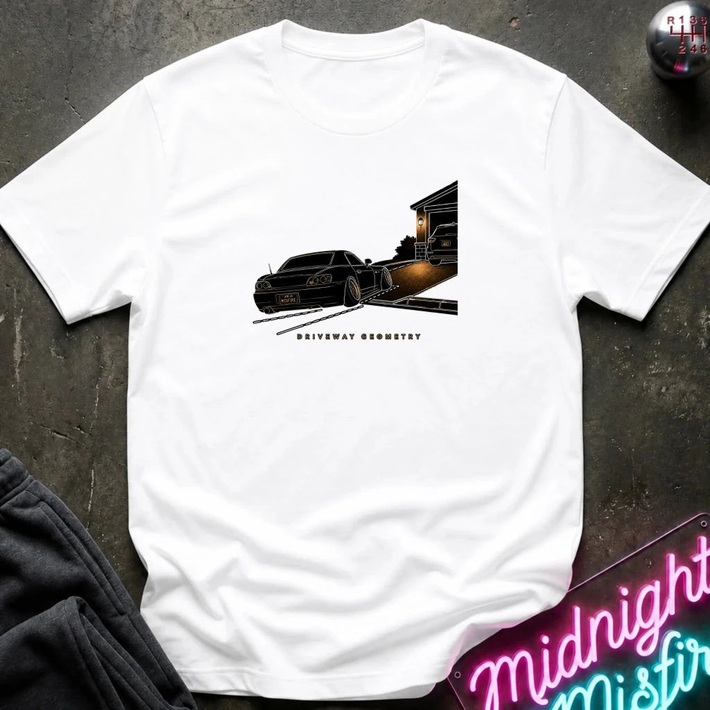 A white short-sleeve t-shirt laid flat on a dark textured surface, featuring a graphic print of a black classic car going up the driveway.