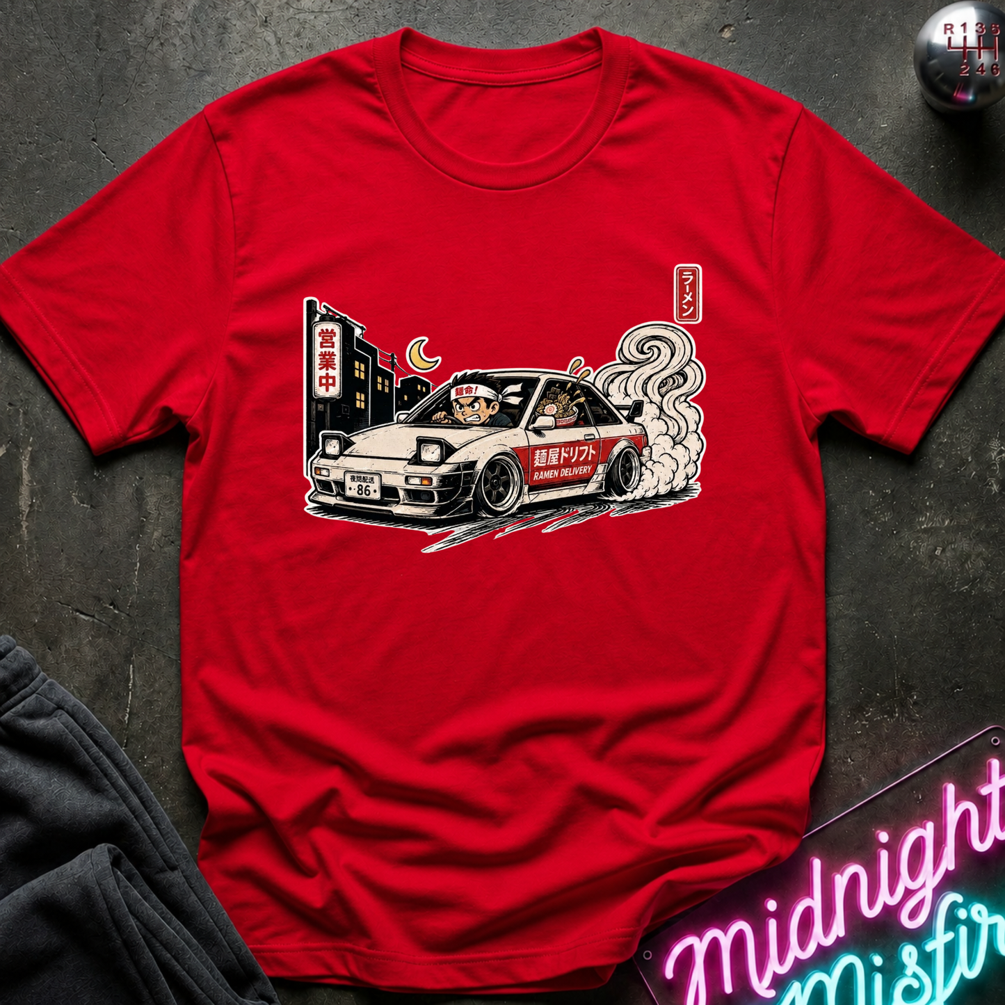 A red short-sleeve graphic T-shirt laid flat on a dark textured surface, featuring a cartoon-style car illustration with smoke, Japanese signage, and a crescent moon. Partially visible props include dark fabric at lower left and a neon-style sign at lower right.