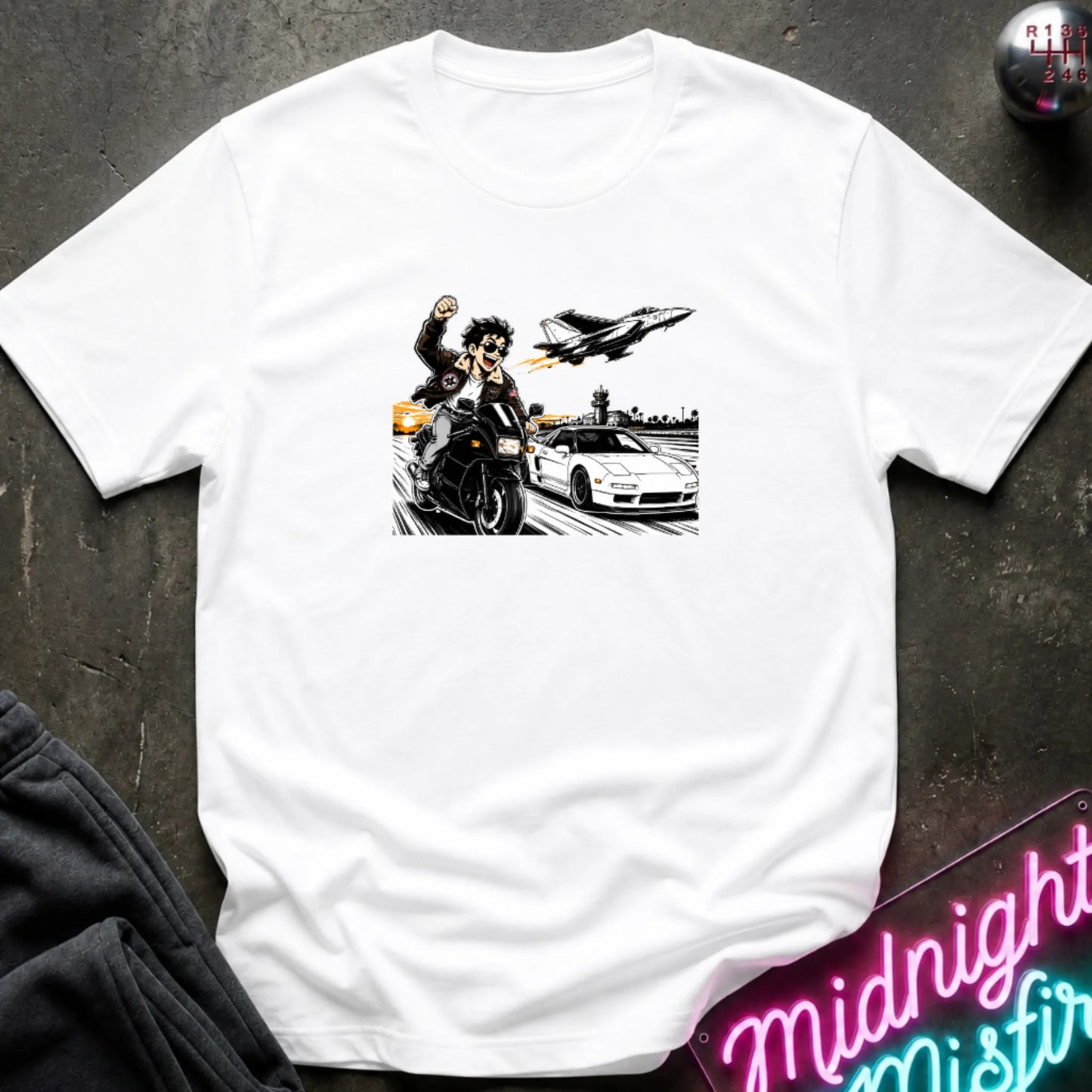 A white short-sleeve crewneck t-shirt with a graphic print of a motorcycle racer, a sports car, and a jet plane, laid flat on a dark textured background with a neon sign and sweatpants partially visible.
