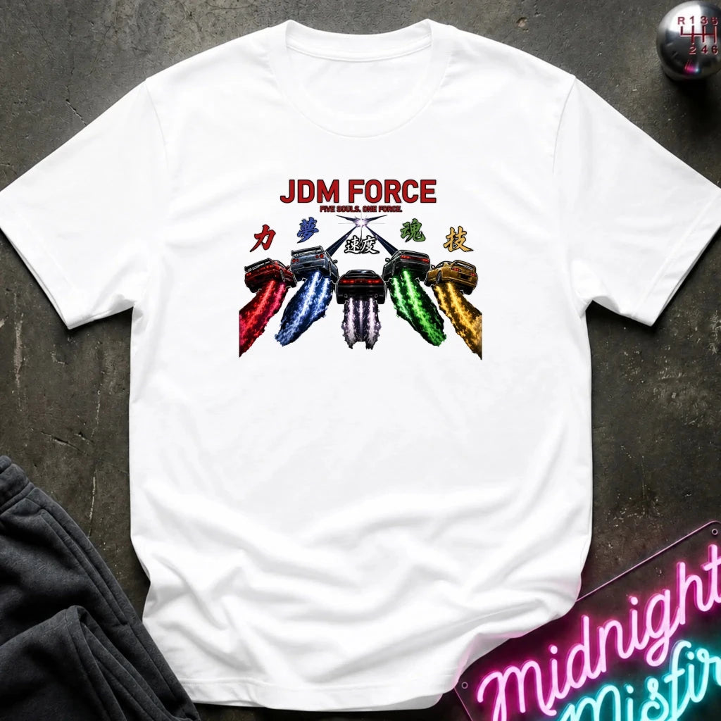 A white short-sleeve graphic T-shirt laid flat on a dark textured surface. The shirt features a colorful graphic of five stylized car headlights in red, blue, green, white, and gold, with the text 'JDM FORCE' in red above them.