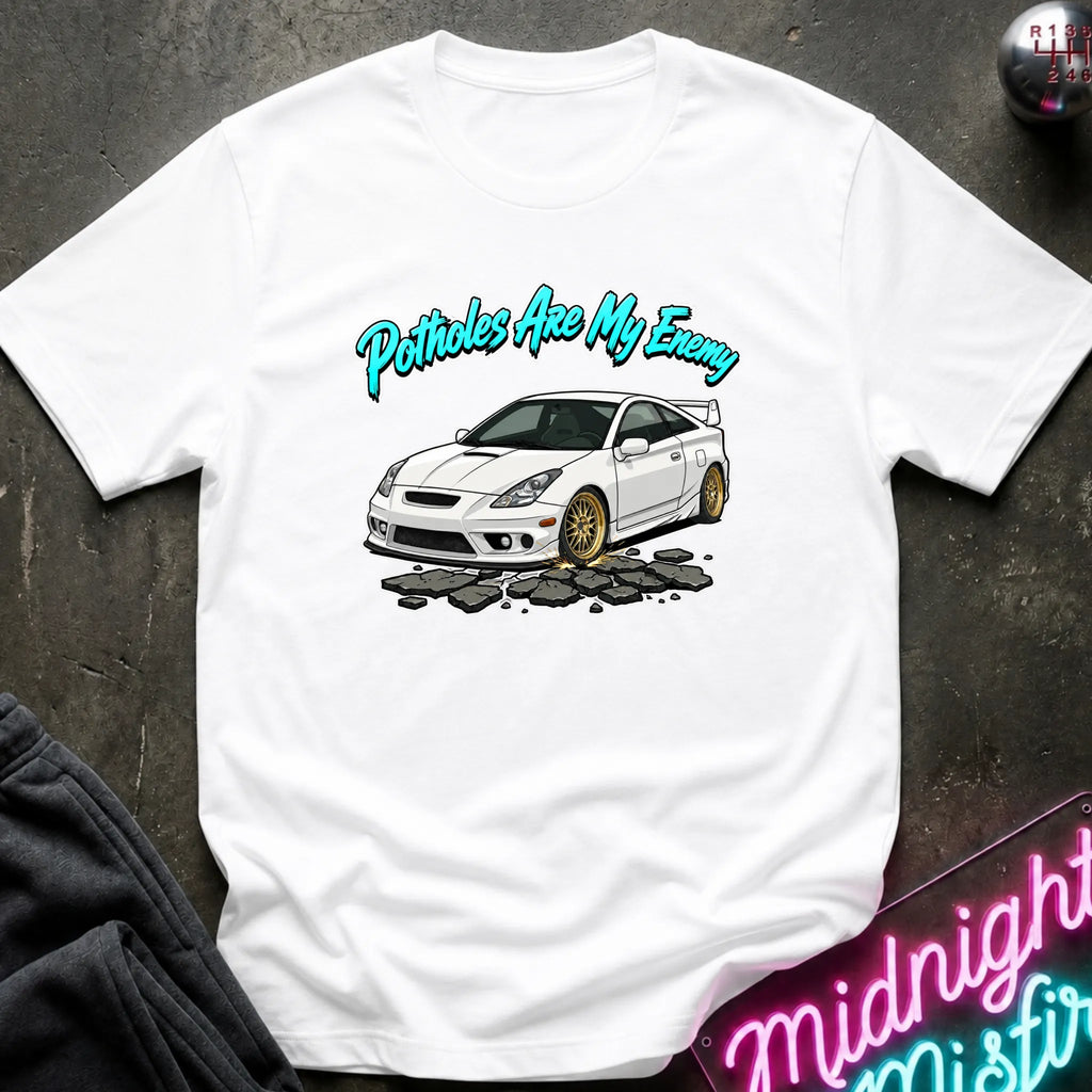 A white short-sleeve graphic T-shirt laid flat on a dark textured surface. The shirt features a printed illustration of a white sports car with gold wheels and the text 'Potholes Are My Enemy' in bright teal script above it.