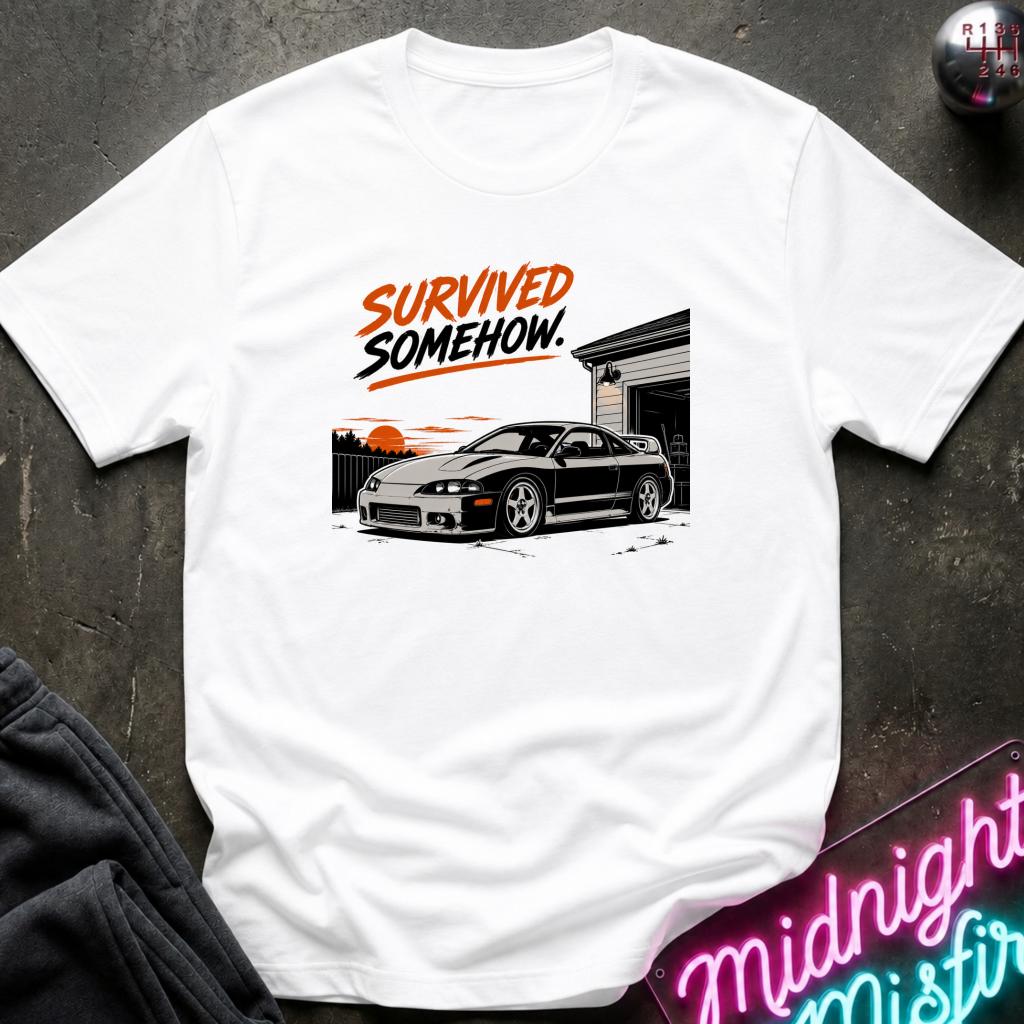 A white short-sleeve graphic T-shirt laid flat on a dark textured surface. The shirt features a printed design of a black sports car in front of a garage with the text 'SURVIVED SOMEHOW.