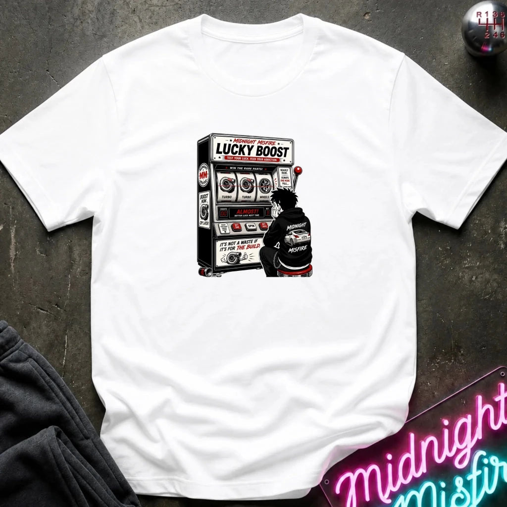 A white short-sleeve crewneck t-shirt with a graphic print of a person playing a slot machine labeled 'LUCKY BOOST'. The shirt is displayed flat on a dark textured surface with a neon sign and dark pants partially visible in the corners.