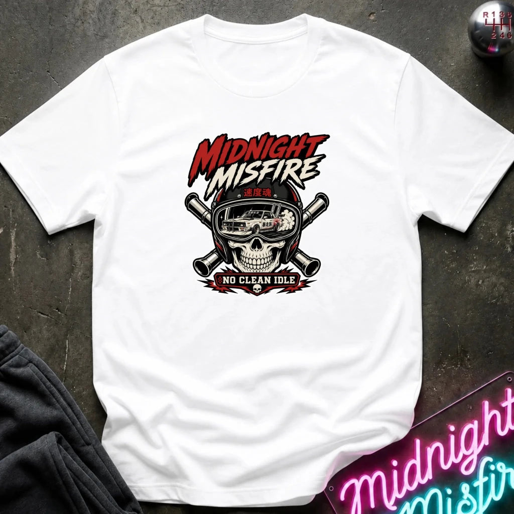 A white graphic t-shirt laid flat on a dark textured surface. The shirt features a central graphic of a skull wearing a racing helmet with crossed pipes behind it, and the text 'MIDNIGHT MISFIRE' and 'NO CLEAN IDLE'.