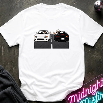 A white short-sleeve t-shirt laid flat on a dark textured surface, featuring a graphic print of a white convertible sports car facing a black sports car in a split scene. Styled flat-lay props include a dark sweatshirt in the lower-left corner and a neon sign in the lower-right.