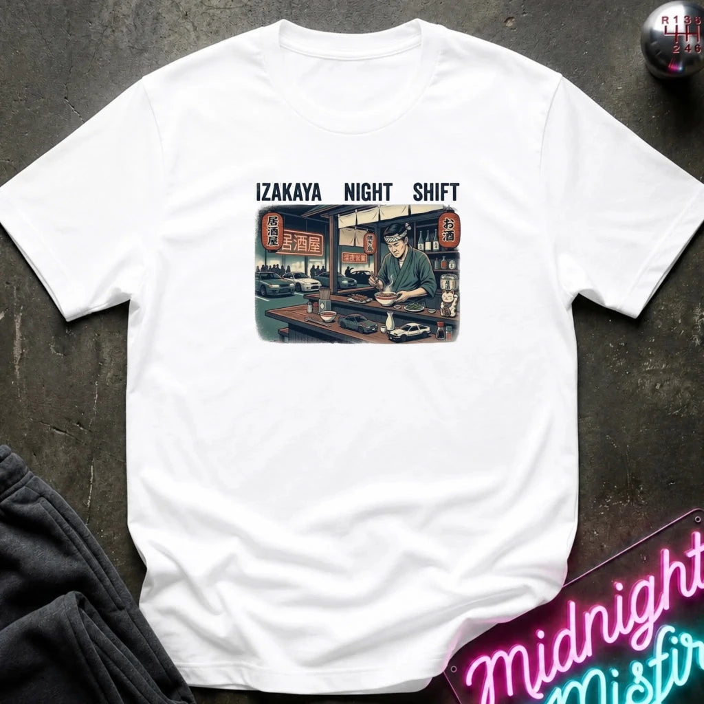 A white short-sleeve t-shirt laid flat on a dark textured surface, featuring a vintage-style graphic of an izakaya scene with the text 'IZAKAYA NIGHT SHIFT' above it. 