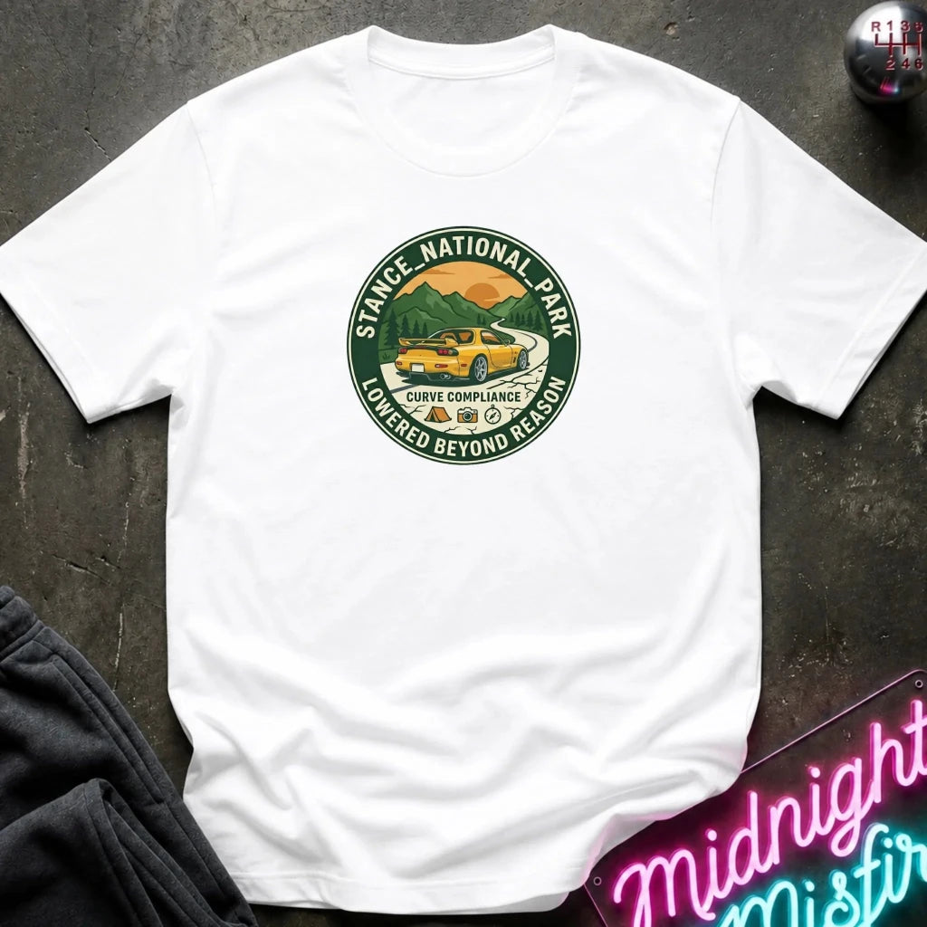 A white short-sleeve crewneck t-shirt laid flat on a dark textured surface. The shirt features a circular graphic in the center depicting a yellow taxi driving through a mountain landscape, 