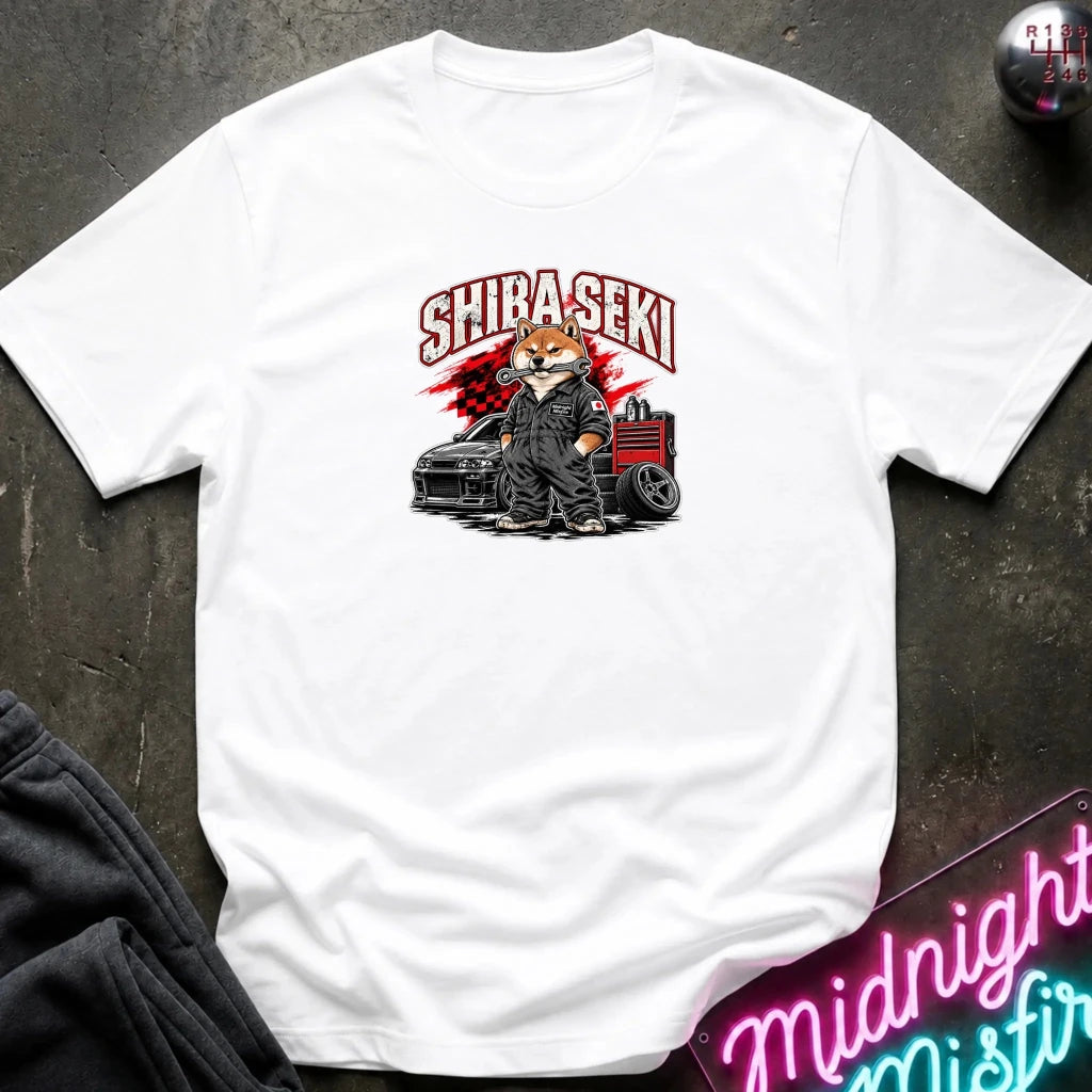 A white short-sleeve graphic T-shirt laid flat on a dark textured surface. The shirt features a stylized illustration of a Shiba Inu character in a black suit standing next to a black car and red tool chest, with the word 'SHIBA SEKI' printed above in red and white.