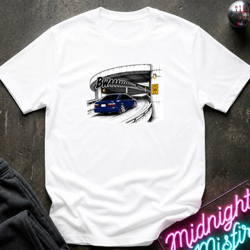 A white short-sleeve crewneck t-shirt laid flat on a dark textured surface. The shirt features a graphic print of a blue car driving under a highway overpass with the text 'BWAHAHA'.