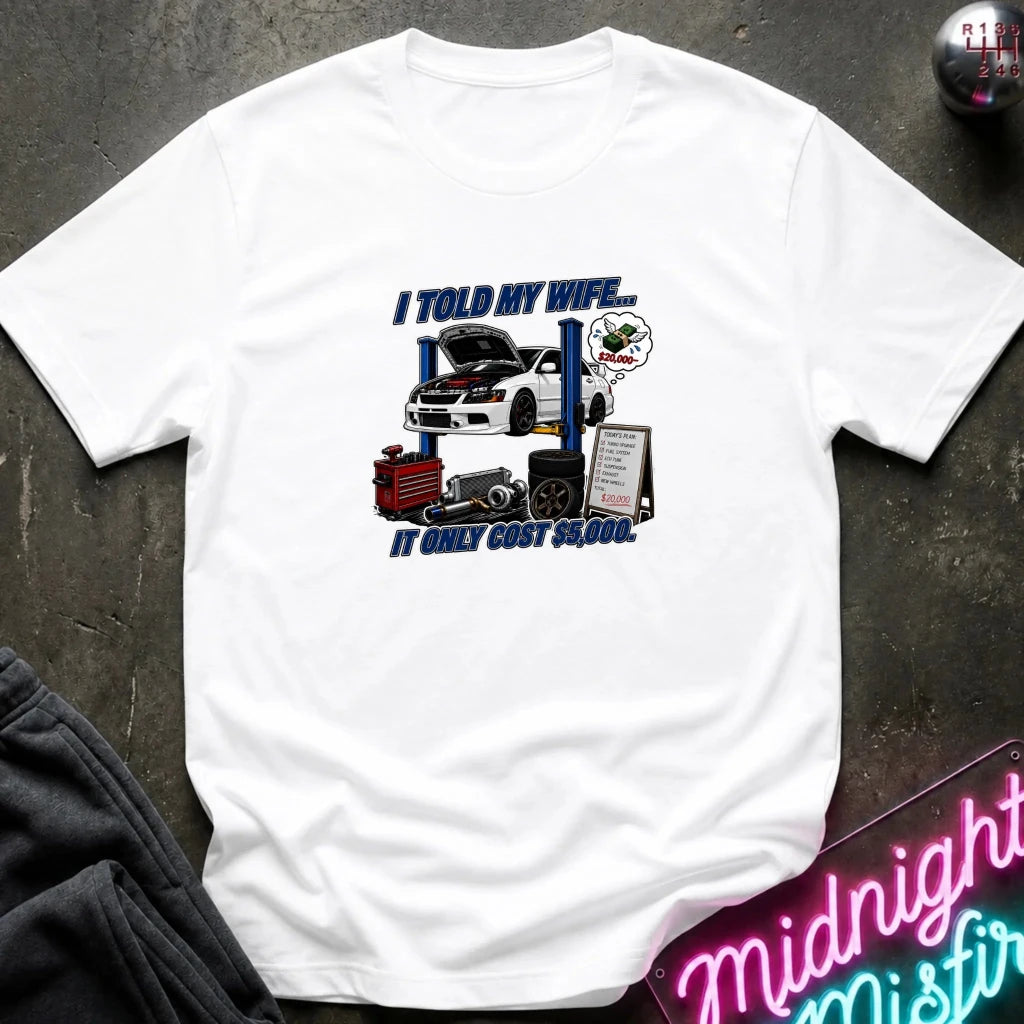A white short-sleeve graphic t-shirt laid flat on a dark textured surface. The graphic features a white sports car on a blue car lift with the text 'I TOLD MY WIFE.