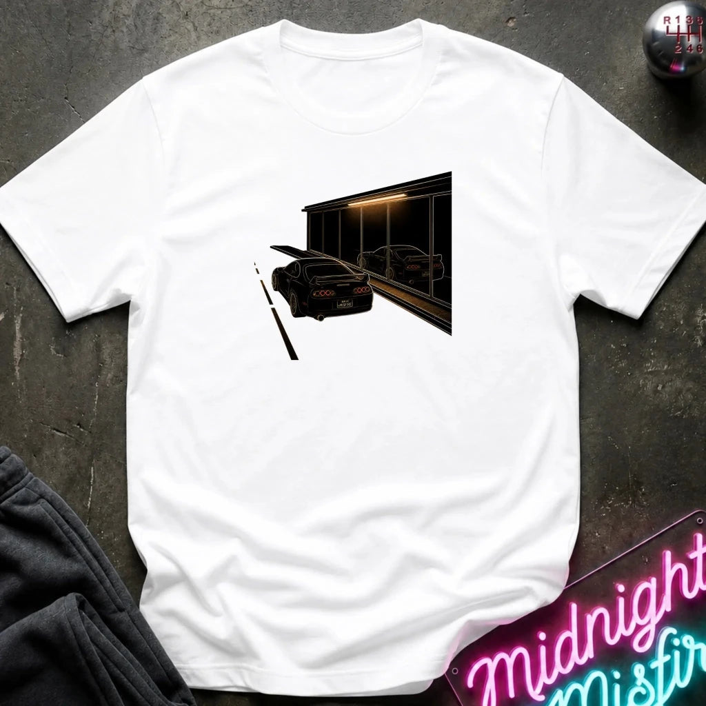 A white short-sleeve t-shirt laid flat on a dark textured surface, featuring a graphic print of a black sports car in front of a dark building with warm light. 