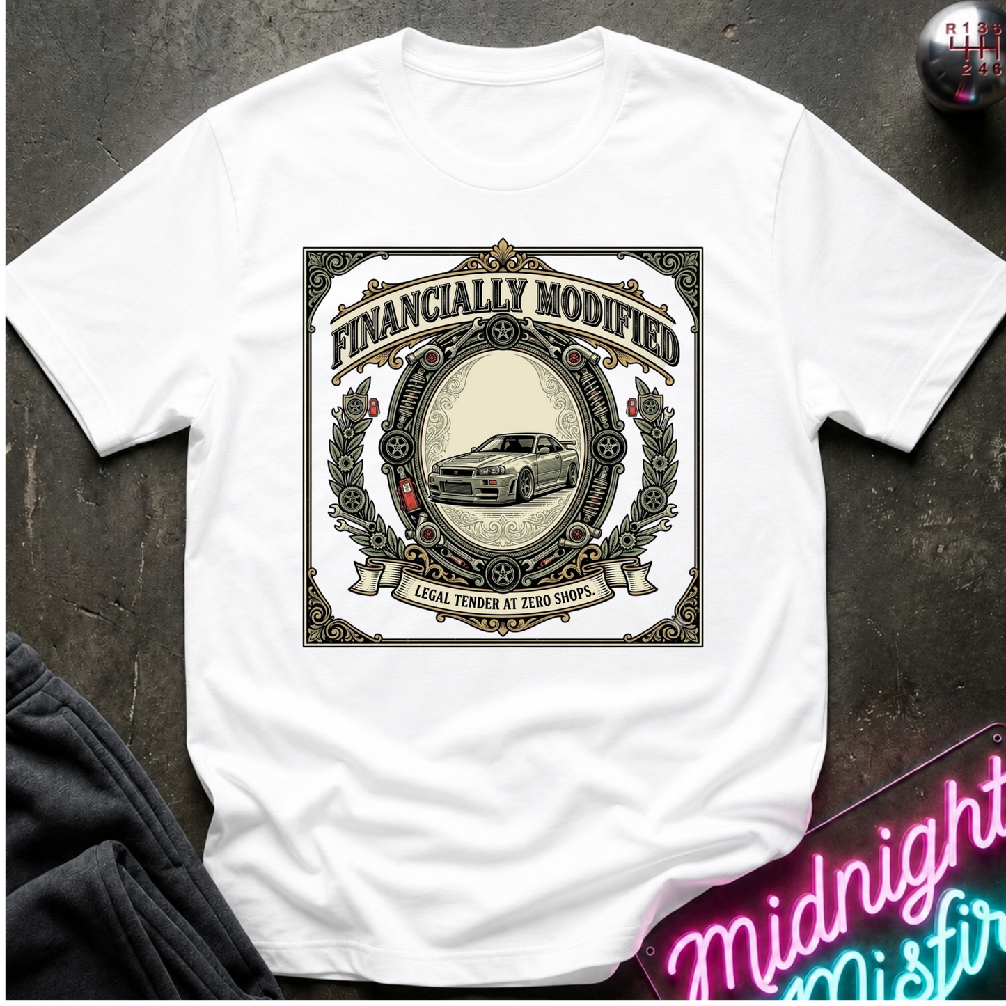 A white short-sleeve graphic t-shirt laid flat on a dark textured surface. The shirt features a large vintage-style circular graphic with the text 'FINANCALLY MODIFIED' and a car illustration.