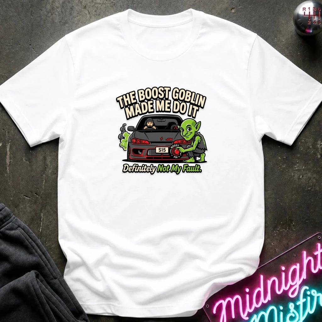 A white short-sleeve graphic t-shirt laid flat on a dark textured surface. The graphic features a green cartoon character next to a red sports car with the text 'THE BOOST GOBLIN MADE ME DO IT' above and 'Definitely Not My Fault.