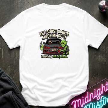 A white short-sleeve graphic t-shirt laid flat on a dark textured surface. The graphic features a green cartoon character next to a red sports car with the text 'THE BOOST GOBLIN MADE ME DO IT' above and 'Definitely Not My Fault.