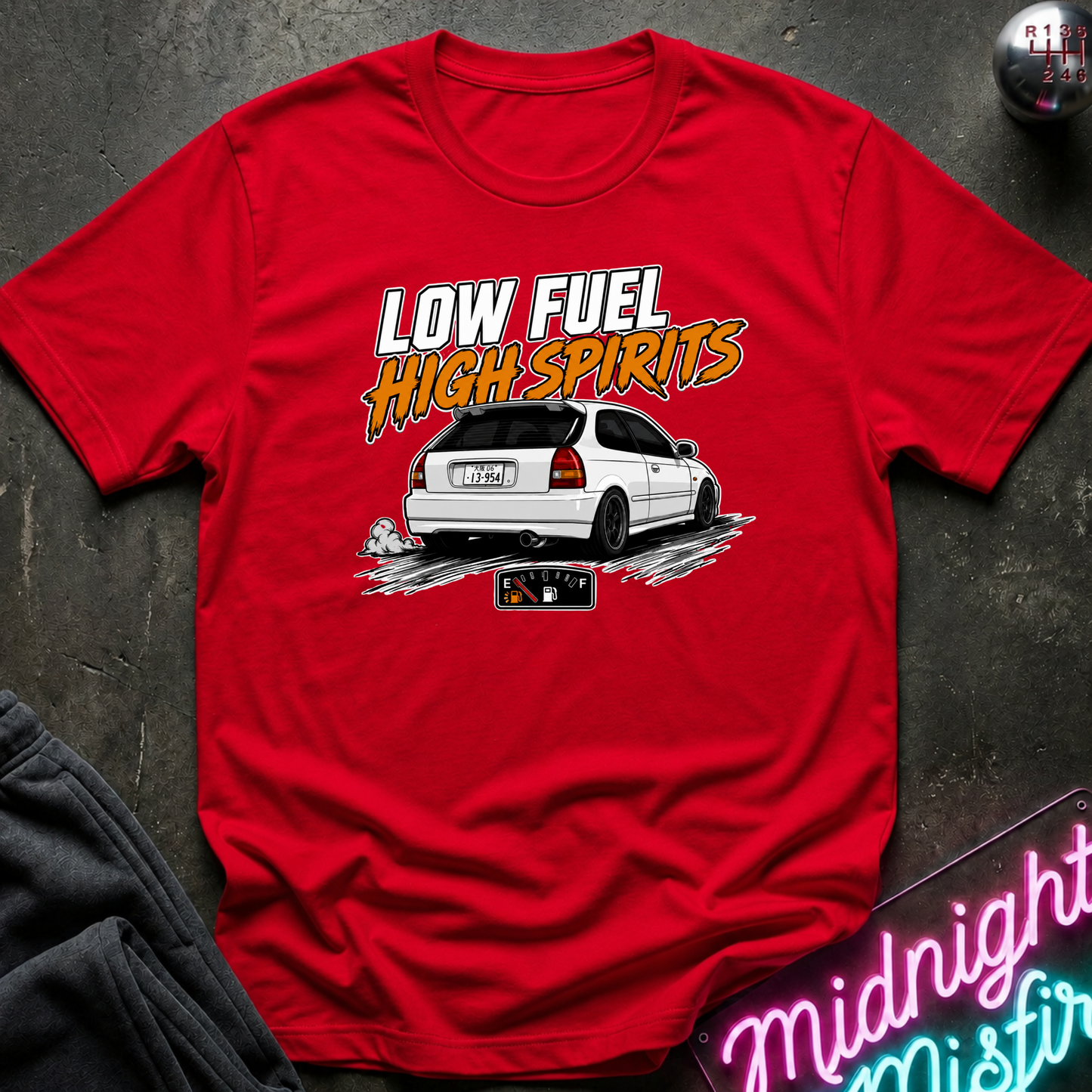 A red short-sleeve graphic T-shirt laid flat on a dark textured surface. The shirt features a white car illustration and the text 'LOW FUEL HIGH SPIRITS' in white and orange.