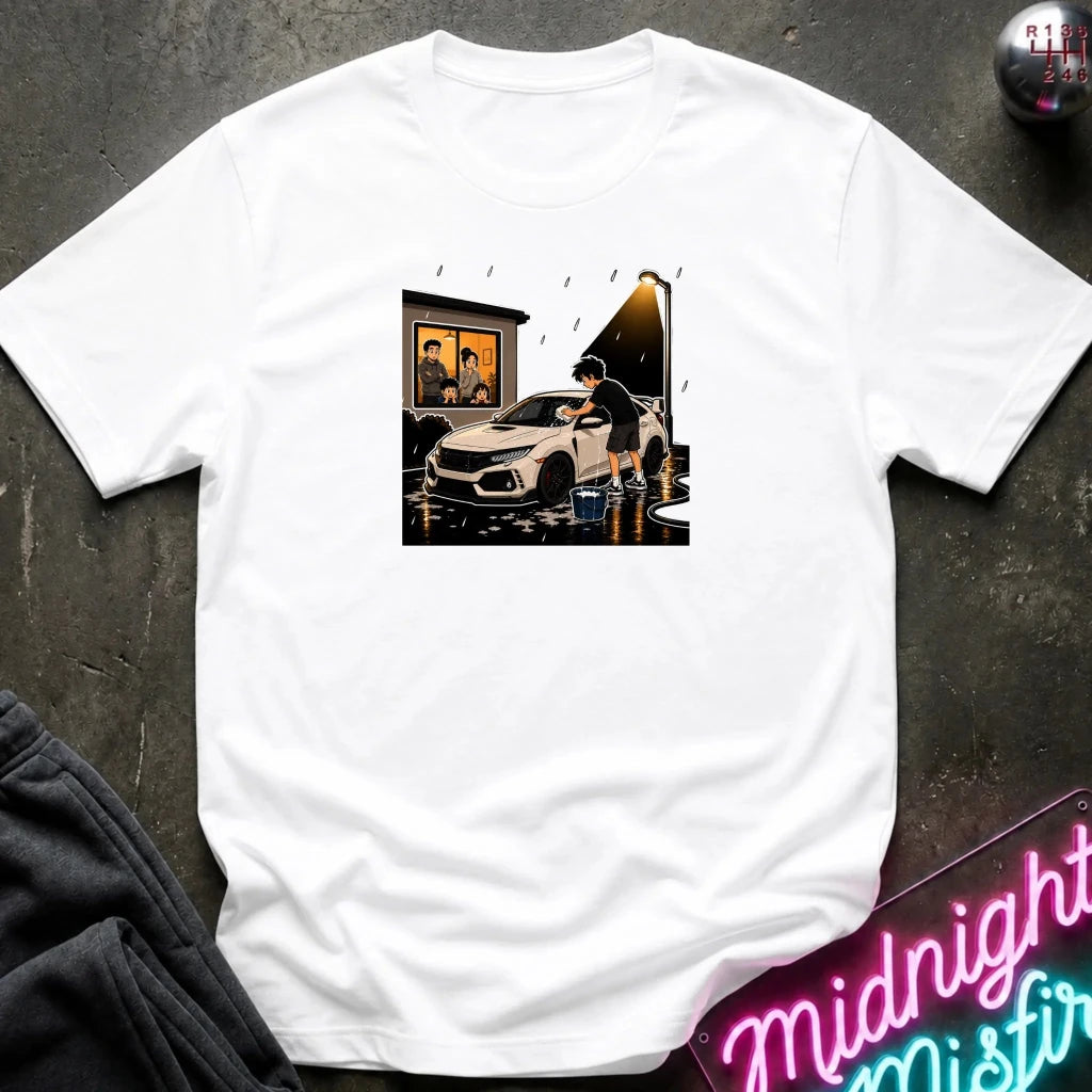A white short-sleeve crewneck t-shirt laid flat on a dark textured surface, featuring a central graphic print of a white sports car in a rainy street scene. 