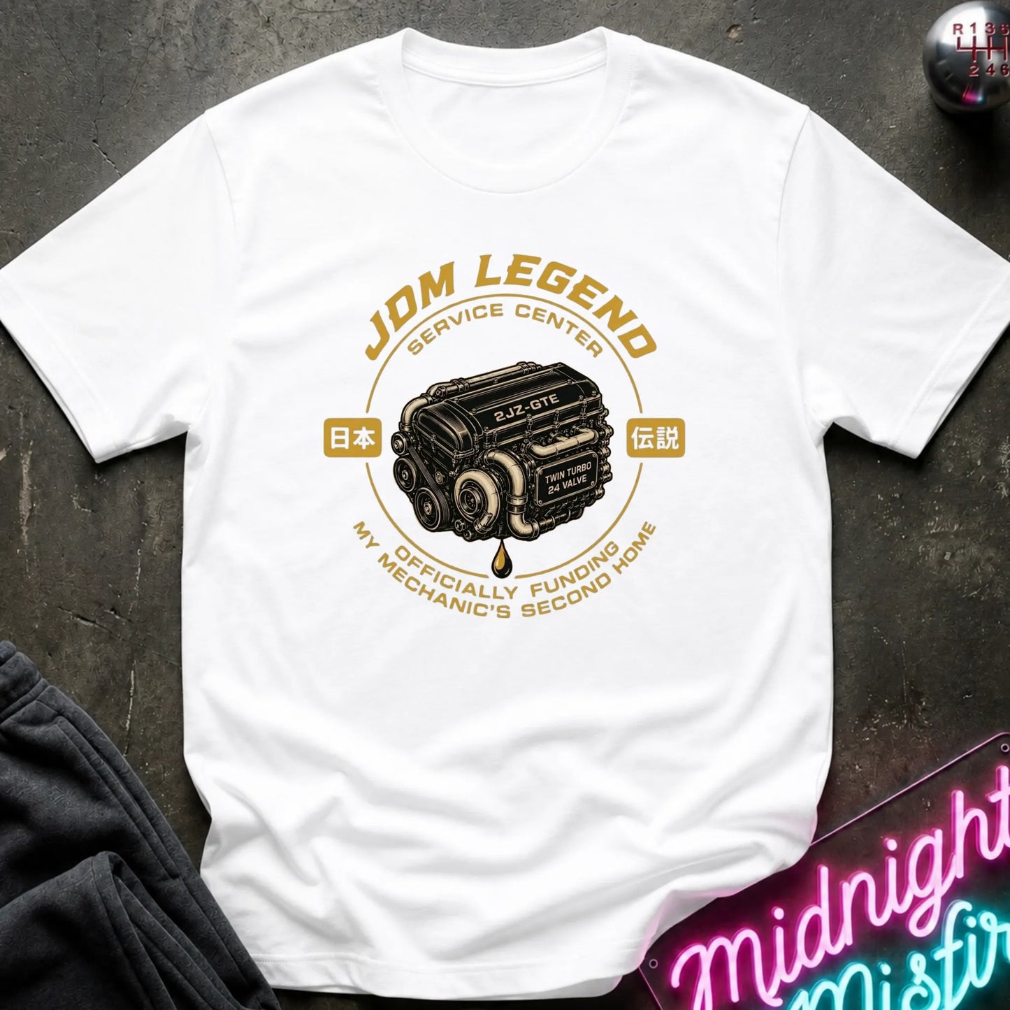 A white short-sleeve graphic T-shirt laid flat on a dark textured surface. The shirt features a vintage-style circular print with a black and gold Japanese car illustration and the text 'JDM LEGEND SERVICE CENTER' and 'MY OFFICIALLY FUNDING MY MECHANIC'S SECOND HOME.