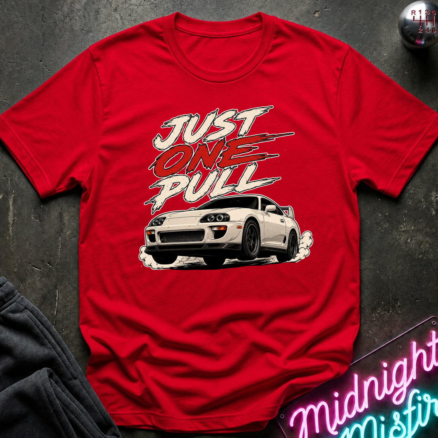 A red short-sleeve graphic T-shirt laid flat on a dark textured surface. The shirt features a white sports car and the words 'JUST ONE PULL' in stylized white and red lettering.