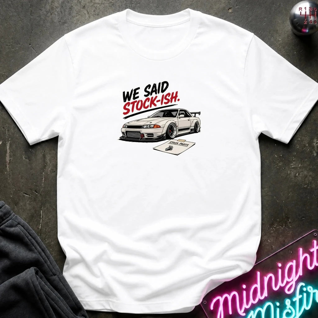 A white short-sleeve t-shirt laid flat on a dark textured surface, featuring a graphic print of a pink convertible car in front of a building with the text 'SAKURA MOTORS' and cherry blossoms. 