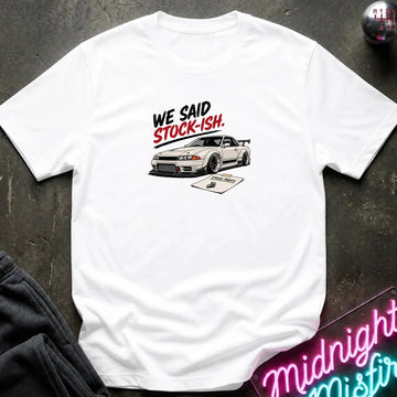 A white short-sleeve t-shirt laid flat on a dark textured surface, featuring a graphic print of a pink convertible car in front of a building with the text 'SAKURA MOTORS' and cherry blossoms. 