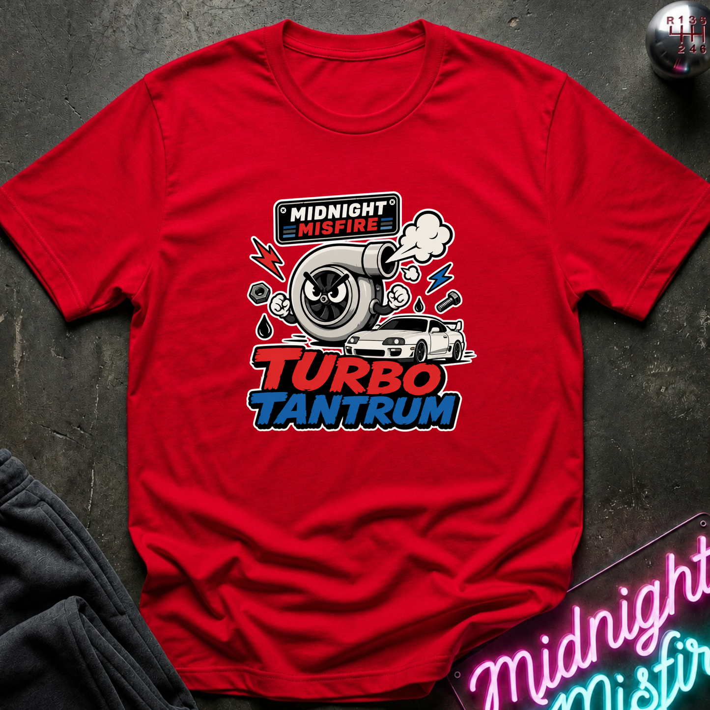 A red short-sleeve graphic T-shirt laid flat on a dark textured surface. The shirt features a central turbo-themed print with the words 'MIDNIGHT MISFIRE' and 'TURBO TANTRUM' in red, white, and blue, plus a cartoon turbocharger, car, smoke, and lightning accents.