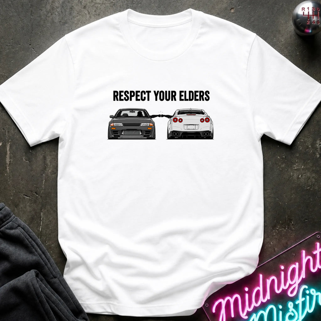 A white short-sleeve graphic T-shirt laid flat on a dark textured surface. The shirt has a black printed slogan reading 'RESPECT YOUR ELDERS' above two cars, with a neon sign and dark clothing partially visible around it.
