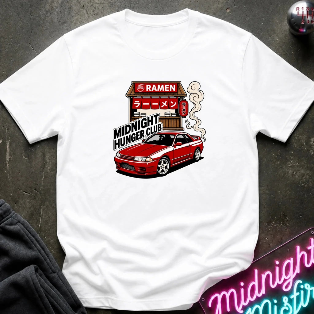 A white short-sleeve graphic t-shirt featuring a red Nissan Sentra car and a ramen shop illustration with the text 'MIDNIGHT HUNGER CLUB'. The shirt is displayed flat on a dark textured surface with a neon sign and dark sweatpants partially visible.