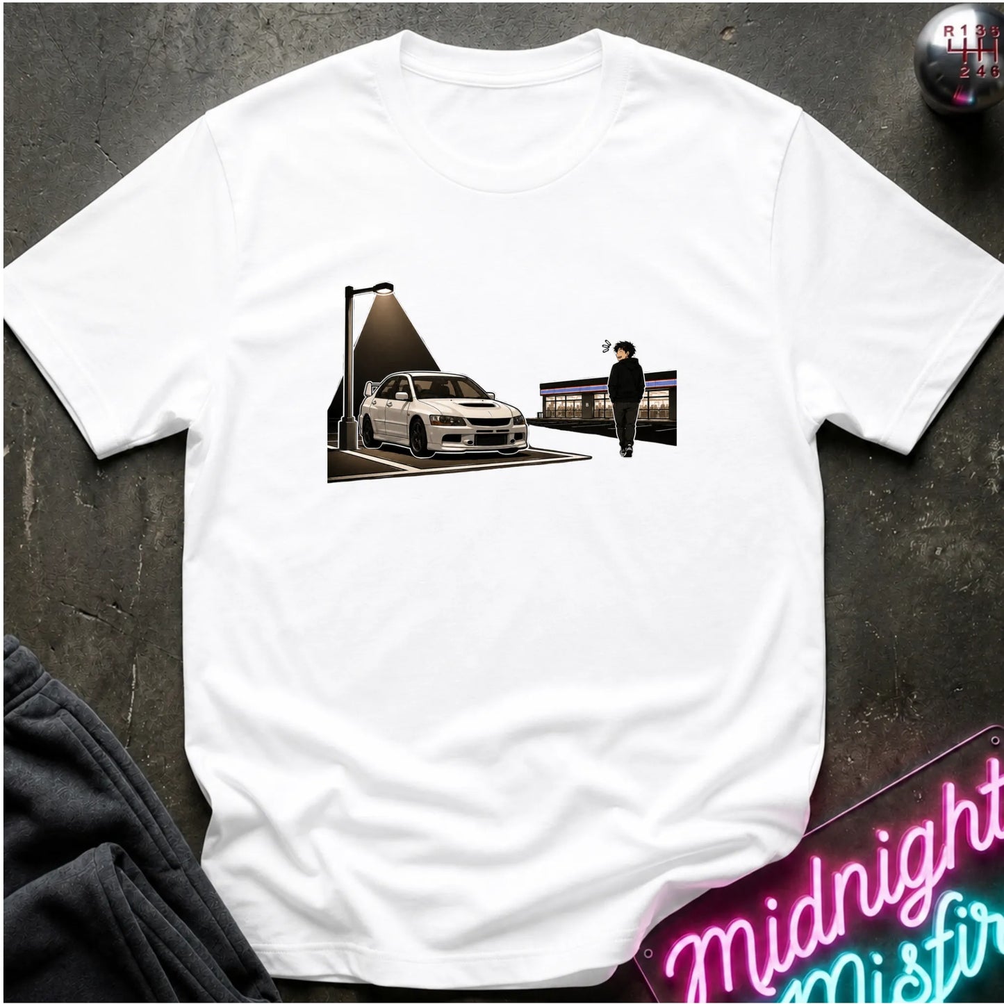 A white short-sleeve t-shirt laid flat on a dark textured surface, featuring a rectangular graphic print on the chest of a white sports car under a streetlamp with a walking figure in the foreground. A neon sign is partially visible in the bottom right corner.