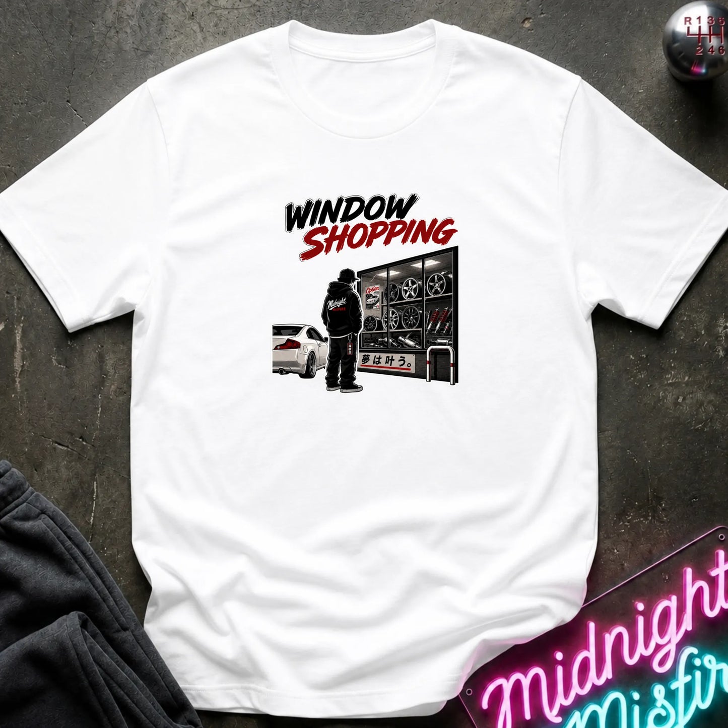 A white short-sleeved t-shirt  that has a cars parked and a person checking out windows of parts.