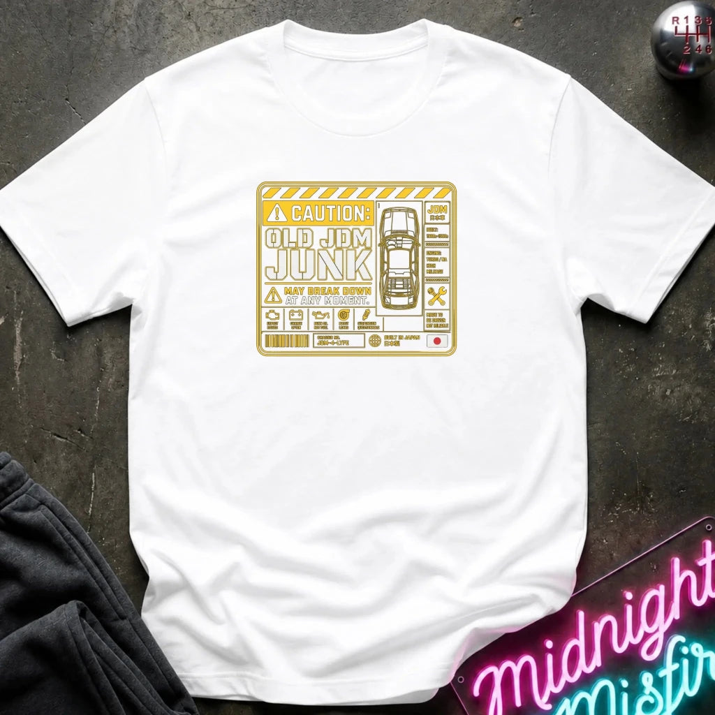 A white short-sleeve crewneck t-shirt featuring a vintage-style yellow and orange graphic of a car and the text 'CAUTION: OLD JdM JUNK'. The shirt is displayed flat on a dark textured surface with a neon sign and sweatpants partially visible.
