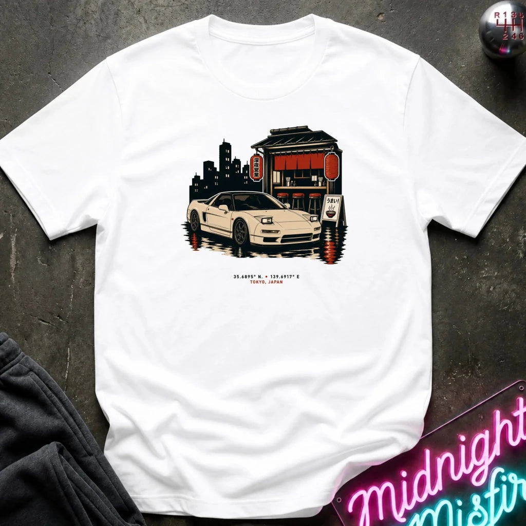 A white short-sleeve t-shirt laid flat on a dark textured surface, featuring a graphic print of a white sports car parked in front of a red and black building with a city skyline behind it. Small red text is printed below the graphic.