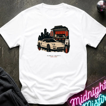 A white short-sleeve t-shirt laid flat on a dark textured surface, featuring a graphic print of a white sports car parked in front of a red and black building with a city skyline behind it. Small red text is printed below the graphic.